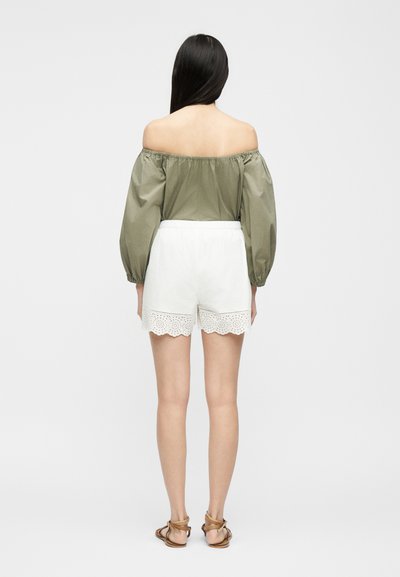 Γυναίκα που στέκεται με την πλάτη να φαίνεται, φοράει μακρυμάνικη μπλούζα σε ελιά με ακάλυπτους τους ώμους, λευκά σορτς από δαντέλα eyelet και καφέ σανδάλια.