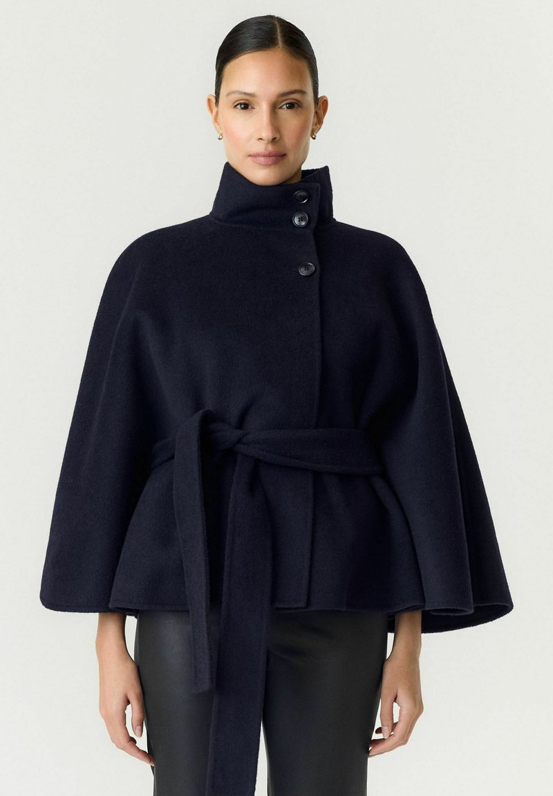 Femme aux cheveux tirés en arrière porte un manteau cape bleu marine foncé boutonné avec un col haut et une ceinture à la taille sur un pantalon en cuir.