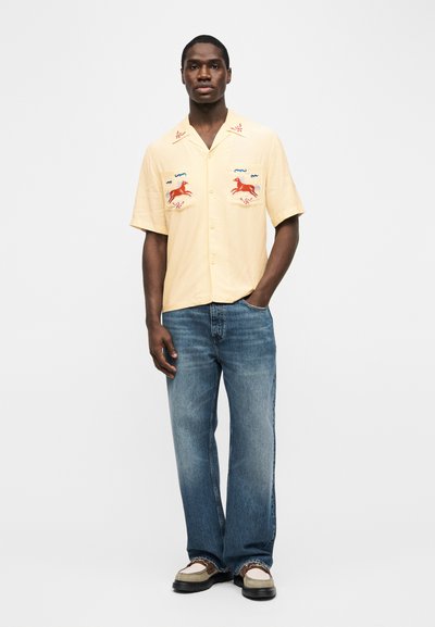 Άνδρας μοντέλο που φοράει ανοιχτό κίτρινο πουκάμισο με κοντό μανίκι και κεντητό κόκκινο άλογο, μπλε τζιν και δίχρωμα loafers, στέκεται με το ένα χέρι στην τσέπη.