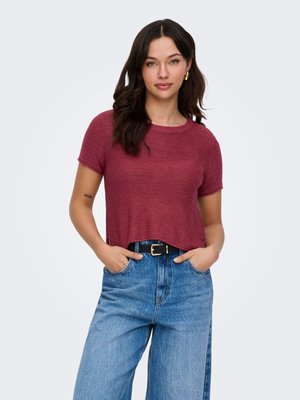 Jeune femme aux cheveux bruns ondulés portant un haut en maille bordeaux à manches courtes et un jean taille haute bleu avec ceinture noire, les mains dans les poches.