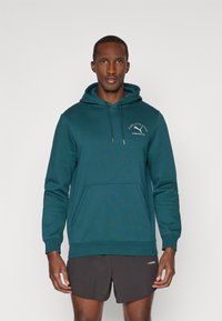 Sudadera con capucha en color teal, con bolsillo frontal, cordón y logo bordado. La tela parece suave y texturizada. El modelo lleva puestos unos pantalones cortos negros.