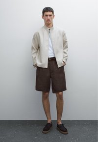 Veste légère beige avec un col montant et un seul bouton, associée à un short en lin marron et des chaussures en daim foncé. Éléments de design simples.
