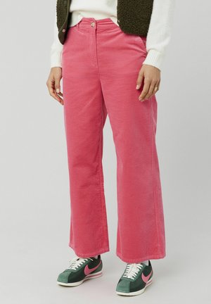 Pantalons en velours côtelé rose avec des jambes larges et des rayures texturées. Portés avec des baskets vertes et roses, dotés d'une fermeture à boutons et de poches.