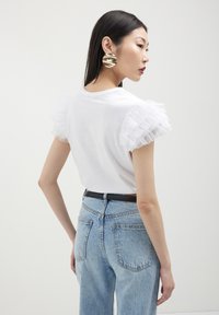 Donna che indossa una camicia bianca con maniche arricciate, jeans blu con una cintura nera e grandi orecchini rotondi dorati, girata di lato su uno sfondo semplice.