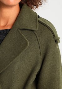 Olivgrön ullkappa med breda slag och knappdetalj; tyget verkar mjukt med en texturerad yta. Närbild på axel och krage.