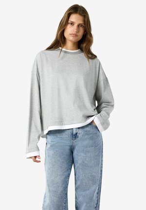Joven mujer con pelo largo y castaño que lleva una sudadera gris suelta sobre una camisa blanca y vaqueros azul claro sobre un fondo liso.