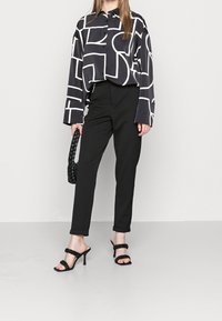 Camisa negra de manga larga con patrón geométrico blanco, pantalones negros ajustados y sandalias de tacón negras con tiras, llevando un bolso trenzado negro.
