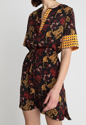 Woman wearing a black romper with yellow and red tiger and floral patterns, featuring yellow geometric trim on sleeves and neckline.