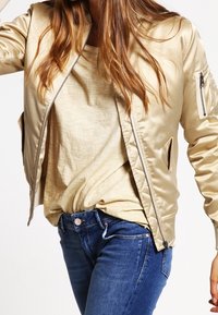 Giacca bomber in satin beige con chiusura a zip, tasche laterali e polsini a costine. Indossata sopra un top beige chiaro e jeans blu.