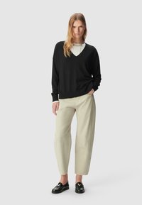 Kobieta ubrana w czarny sweter w serek na białej koszuli, beżowe spodnie, czarne loafersy, stojąca z ręką w kieszeni na jednolitym jasnym tle.