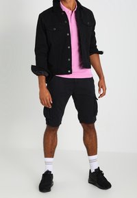 Veste en denim noir avec col en fourrure, polo rose, shorts cargo noirs, chaussettes blanches et baskets noires. Vue de face de la tenue.