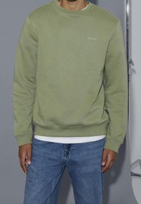 Maglione verde chiaro a girocollo realizzato in tessuto morbido, con polsini e orlo a costine, e un piccolo logo "blend" sul petto. Indossato sopra una camicia bianca.