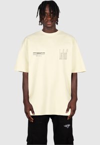 T-shirt a maniche corte di cotone giallo chiaro con elementi grafici di testo; "Gioventù Perduta" stampato in grande font a destra e una frase più piccola a sinistra.
