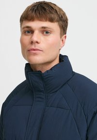 Tamsiai mėlynas pūkinis švarkas su aukštu apykakle, siuvinėjimo tekstūra ir kampuotais siuvinėjimo detalėmis. Lygus medžiaga su lengvu išvaizda.