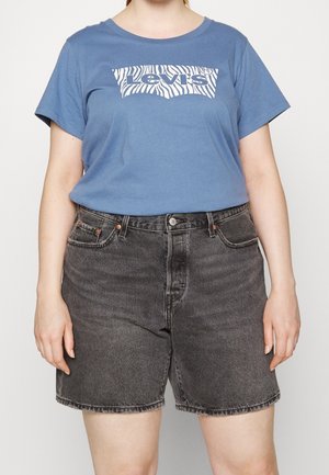 Osoba nosząca niebieski t-shirt Levi's z logo w nadruk zebry i wysokim stanem wyblakłe czarne szorty dżinsowe na jednolitym tle.