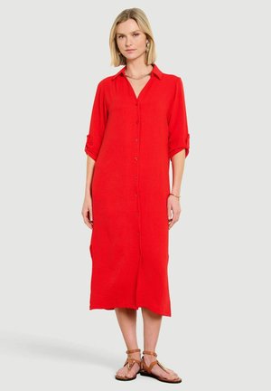 Woman with blonde hair wearing a long red button-up dress with rolled three-quarter sleeves and brown sandals, standing against a plain background.