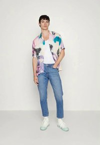 Camisa colorida de manga curta com padrões abstratos em rosa, azul e preto; combinada com jeans ajustados azuis e sapatilhas brancas.