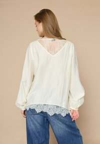 Lyscremebluse med blonde ryg, lange pufærmer og scalloped blondekant, parret med brede blå jeans. Struktureret stof.