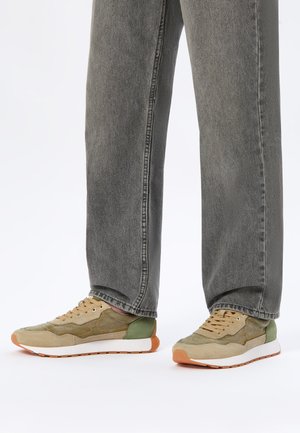 Jambes portant des jeans gris à jambes droites et des baskets décontractées beige, vertes et blanches avec des semelles orange sur fond blanc.