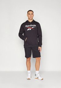 Sudadera negra con capucha con el logo de Reebok, combinada con pantalones cortos negros. Modelo con zapatillas blancas con suelas marrón claro, de pie frente a un fondo neutro.