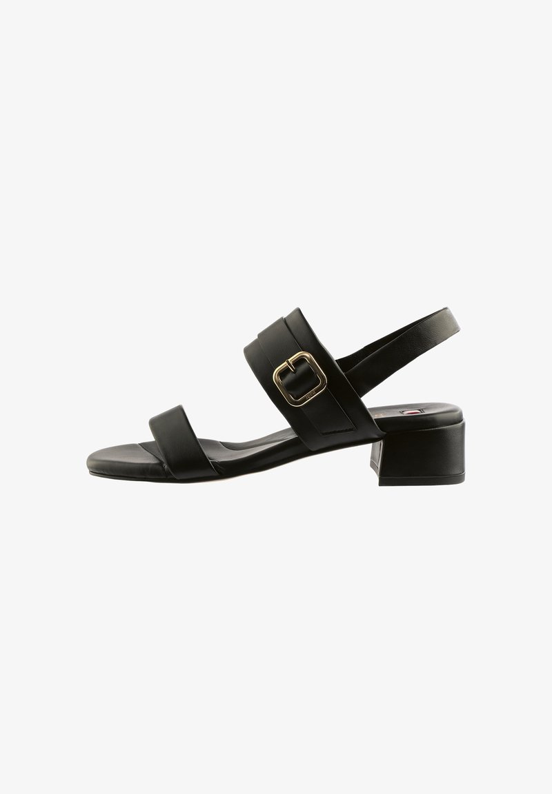 Zwarte sandaal met lage hak en twee bandjes, een gouden gesp op het middelste bandje, en een open achterkant met een hielbandje.
