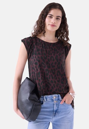 Jeune femme aux cheveux bouclés porte un haut sans manches marron foncé à imprimé léopard, un jean bleu clair, tenant un sac en cuir noir et un bracelet blanc.