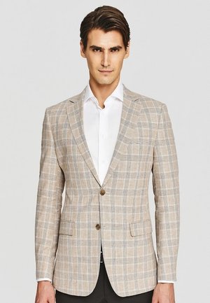 Mężczyzna w beżowej, kratkowanej marynarce na białej koszuli z guzikami, na neutralnym tle, patrzący prosto z neutralnym wyrazem twarzy.