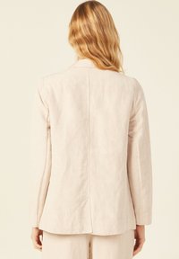 Blazer de lino rosa claro con mangas largas, solapas con muesca y una costura que recorre la parte trasera central. Corte entallado con detalles mínimos.