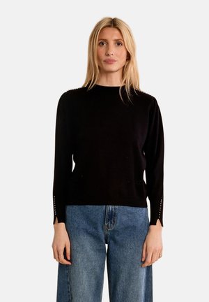 Donna bionda che indossa un top nero a maniche lunghe con dettagli di bottoni sulle maniche e jeans blu a vita alta, in piedi davanti a uno sfondo semplice.