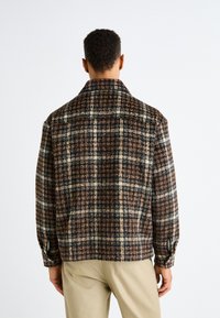 Braun- und cremefarbener Karojacke aus strukturiertem Stoff, ausgestattet mit großem Kragen, knöpfbaren Manschetten und einem lässigen Schnitt. Das Design hebt horizontale und vertikale Linien hervor.