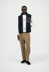 Kék virágmintás polár mellény fekete alapon, cipzáras zsebbel, világos színű hosszú ujjú pólóval és barna kordbársony nadrággal párosítva.