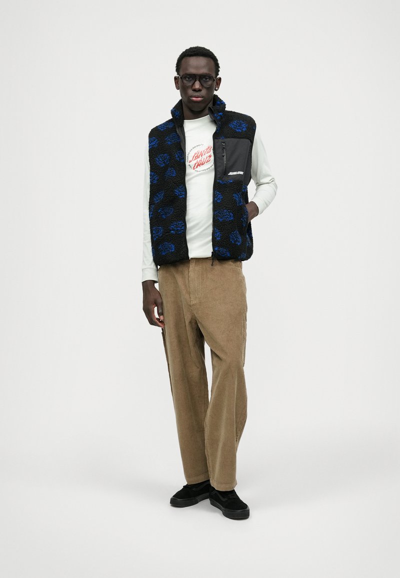 Kék virágmintás polár mellény fekete alapon, cipzáras zsebbel, világos színű hosszú ujjú pólóval és barna kordbársony nadrággal párosítva.