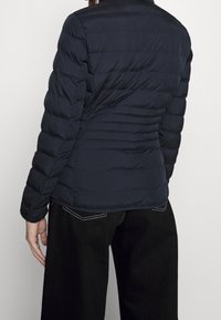 Laivastonsininen toppatakki suoraviivaisella istuvuudella, vaakasuunnassa tikattu kuvio ja pitkät hihat. Ominaisuuksiin kuuluu korkea kaulus ja vähäiset ommelkoristelut.