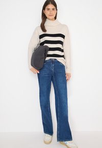Beige und schwarz gestreifter Rollkragenpullover, blaue Schlagjeans, hält eine graue Daunenjacke. Weicher Stoff, lässige Passform und klassische Farben.