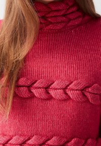 Červený pletený svetr s vysokým vypleteným límcem. Vyznačuje se texturovaným vzorem stehu a výrazným tkaným detailním vrkočem přes hrudník.