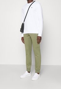 Muž v bílém dlouhém rukávu, olivově zelených teplákových kalhotách s bočními kapsami, bílých teniskách a černé crossbody tašce.