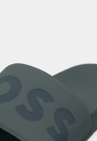 Dark gray slide sandal featuring a textured sole. The strap is smooth with a subtle embossed logo design. Minimalistic and practical shape.
