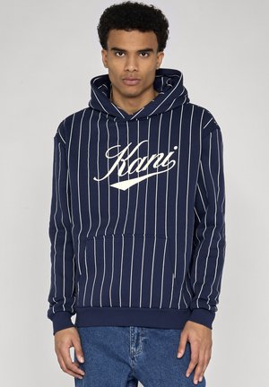 Fiatal férfi sötétkék kapucnis pulóverben, fehér függőleges csíkokkal és "Kani" logóval, kék farmerrel, sima háttér előtt állva.