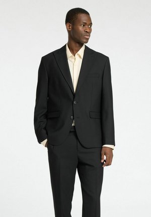 Completo nero su misura con revers a punta, due tasche frontali e pantaloni coordinati. Indossato sopra una camicia leggera beige, tessuto liscio.