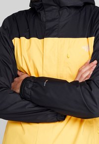 Voděodolná bunda s černobílým designem v černé a žluté barvě, nastavitelnou kapucí a elastickými manžetami pro bezpečné uchycení.