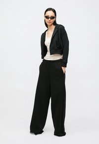 Sweatshirt preto curto com fecho de correr frontal, combinado com calças pretas largas e soltas. Modelo veste um top bege e óculos de sol pretos.