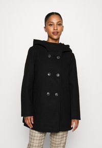 Musta peacoat hupulla, kaksirivinen muotoilu, suuret napit ja pehmeä villamainen tekstuuri. Mukana rento istuvuus ja sivutaskut.