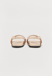Sandales plates beiges avec un design de bride lisse et brillant agrémenté de accents tissés. La semelle est texturée avec un dessous noir pour une meilleure adhérence.