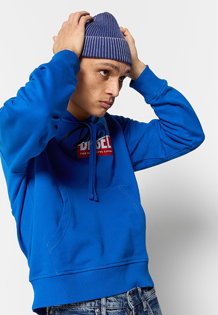 Joven ajustando un gorro de punto morado, vestido con una sudadera Diesel de color azul brillante y jeans, frente a un fondo blanco liso.