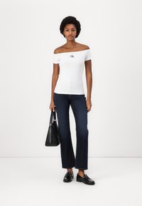 White off-the-shoulder top with a black logo on the chest, paired with dark blue flared jeans and black loafers. Black handbag in hand.