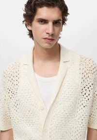 Lichtcrème gebreide cardigan met korte mouwen, met een open haakpatroon en een knoopsluiting aan de voorkant. Draag over een witte tanktop.