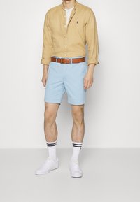 Světle hnědá lněná košile, pod ní bílý tričko, baby modré šortky, hnědý opasek, bílé tenisky, ponožky s černými pruhy. Slim fit, ležérní styl.