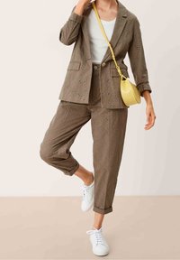 Bruin oogjesstof pak met een elegant gesneden blazer en tapered pants, gecombineerd met witte sneakers en een kleine gele schoudertas.