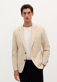 Blazer de mezcla de algodón en color beige con dos botones frontales, solapas de pico y un solo bolsillo en el pecho, llevado sobre una camisa blanca. Textura suave, ajuste entallado.