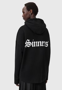 Felpa nera con cappuccio e scritta "Sinners" in bianco, vestibilità rilassata e texture morbida. Il modello la indossa con una gonna nera plissettata.